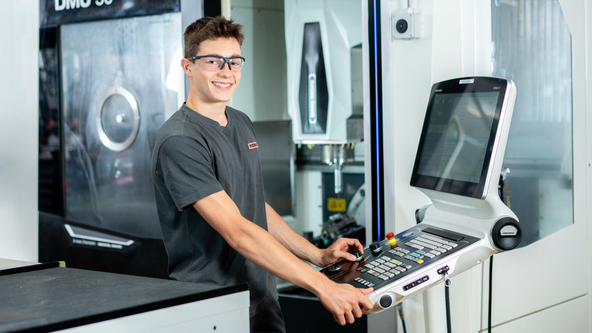 Ein junger Mann mit Schutzbrille und grauem T-Shirt bedient in einer modernen Industrieumgebung ein Bedienfeld und lächelt in die Kamera. Im Hintergrund ist eine große CNC-Maschine zu sehen.