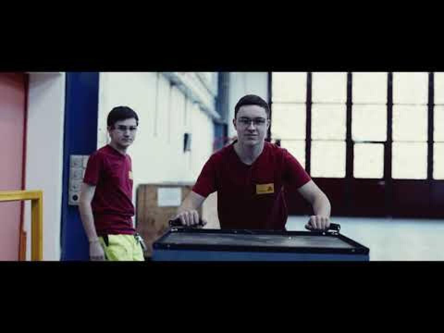 Zwei junge Männer in roten Hemden befinden sich in einer Industrie- oder Lagerhalle. Einer schiebt einen Wagen, der andere steht hinter ihm, beide schauen in die Kamera.