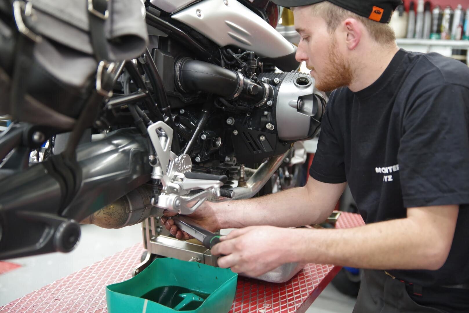 Ein Mechaniker in schwarzem T-Shirt und Mütze arbeitet an einem Motorrad und benutzt einen Schraubenschlüssel im Bereich des Motors. Unter dem Motorrad befindet sich eine grüne Schale, um Flüssigkeiten aufzufangen. Die Szene spielt sich in einer Werkstatt ab.