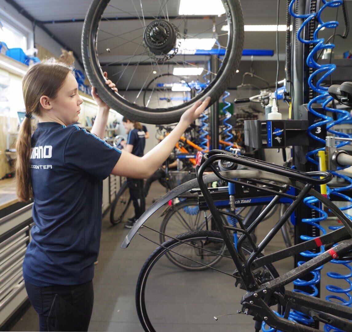 Eine junge Frau hebt in einer Werkstatt ein Fahrradrad in Richtung eines auf einem Reparaturständer montierten Fahrrads, umgeben von Werkzeugen und Geräten in einem gut organisierten Mechanikerarbeitsbereich.