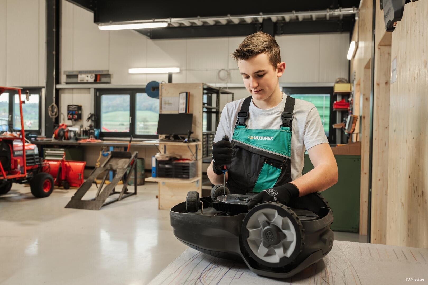 Ein junger Mensch in Overall und Handschuhen repariert einen Roboterrasenmäher in einer Werkstatt mit Werkzeugen, Geräten und einem roten Traktor im Hintergrund.