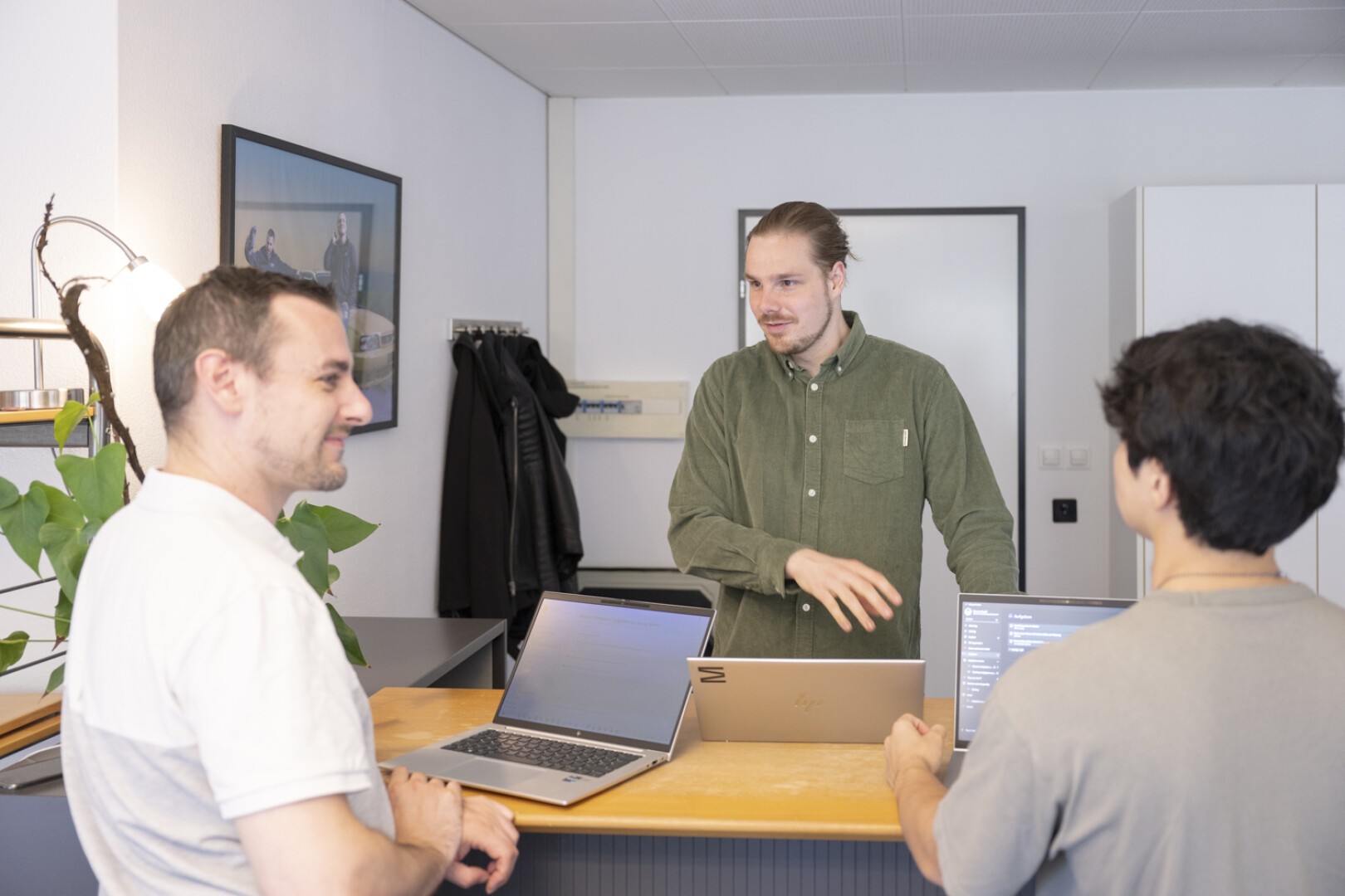 Drei Personen stehen mit Laptops um einen Schreibtisch herum und unterhalten sich in einem hellen Büroraum. Eine Person steht hinter dem Schreibtisch, die anderen stehen auf der gegenüberliegenden Seite.