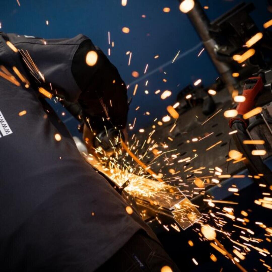 Eine Person, die ein Schutzhemd trägt, bearbeitet einen Metallgegenstand mit einer Schleifmaschine und erzeugt dabei leuchtend orangefarbene Funken vor einem dunkelblauen Hintergrund in einer Werkstattumgebung.