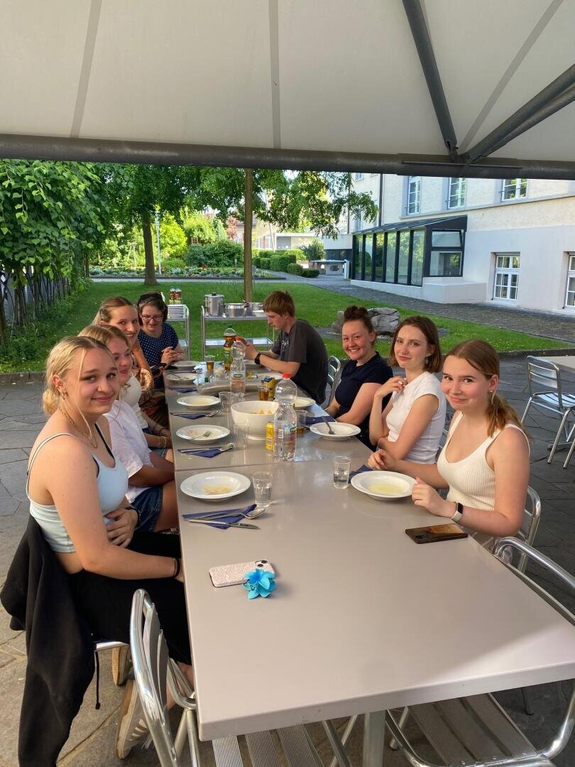 Acht junge Leute sitzen unter einem Sonnendach um einen grauen Tisch im Freien herum, lächeln und unterhalten sich. Auf dem Tisch stehen Teller, Gläser und Getränke. Im Hintergrund sind grüne Bäume und ein Gebäude zu sehen.