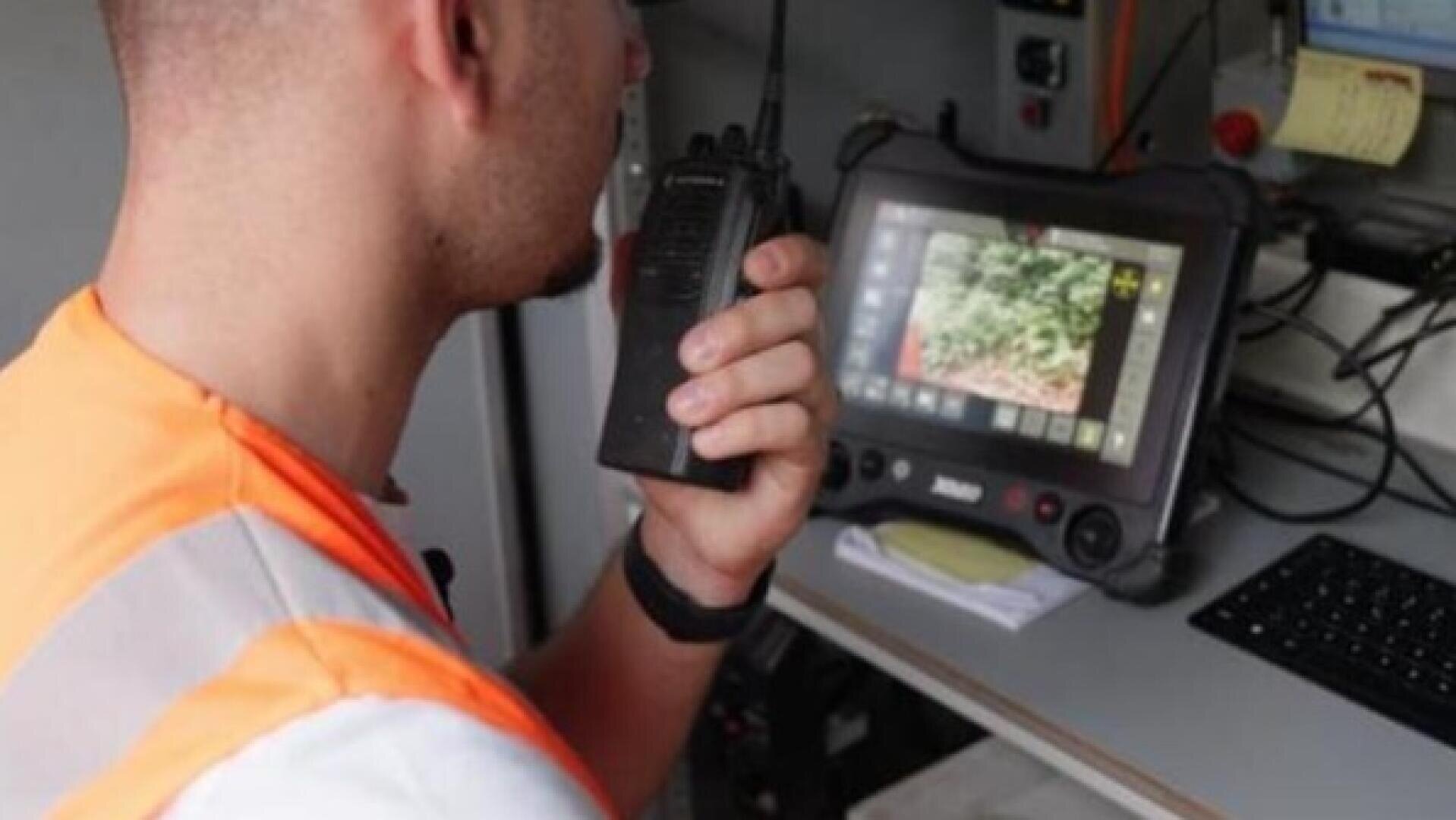 Eine Person in einer Warnweste benutzt ein Walkie-Talkie in der Nähe eines Monitors, auf dem eine Außenaufnahme zu sehen ist; auf dem Schreibtisch liegen eine Tastatur und Notizen.