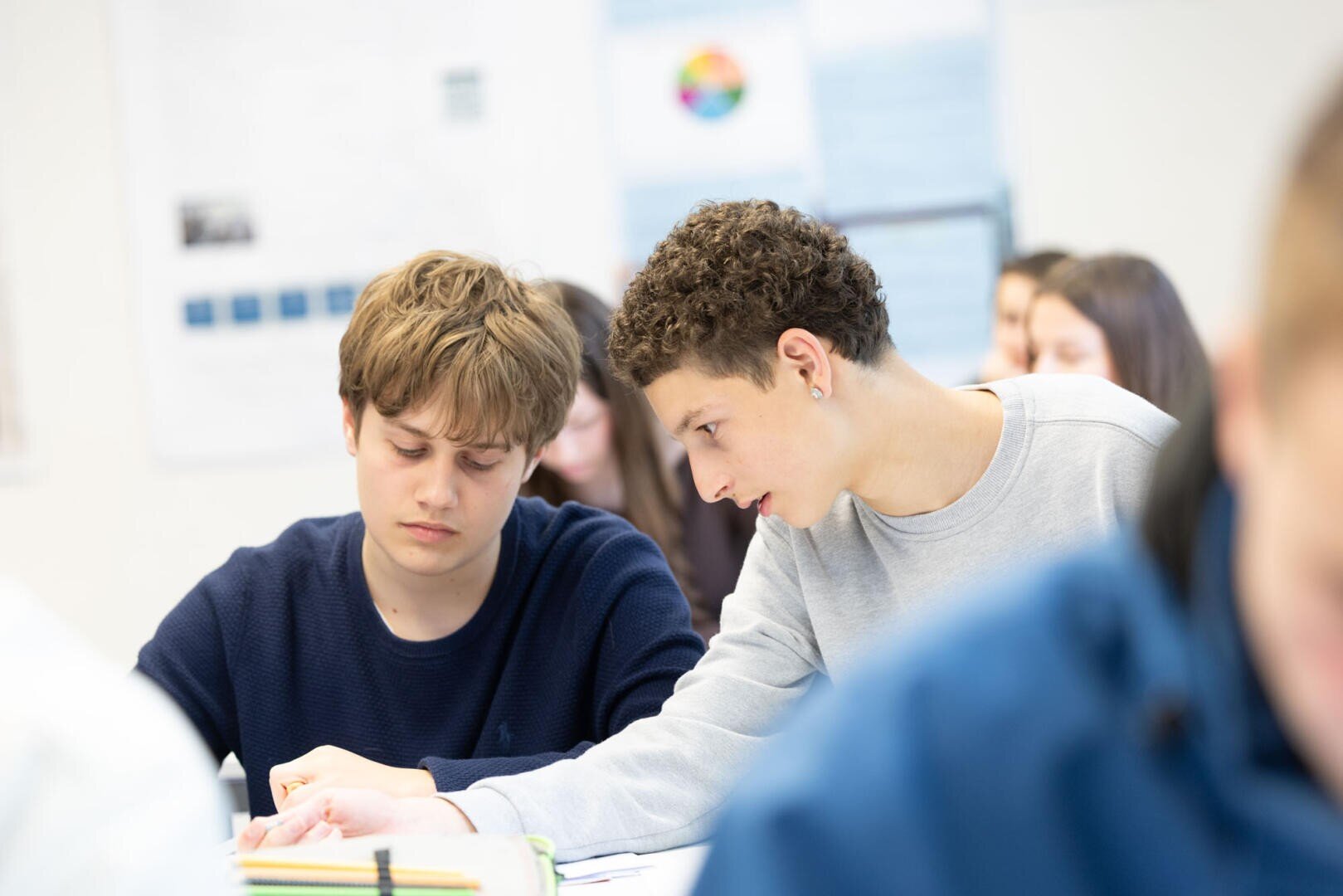 Zwei Schüler sitzen in einem Klassenzimmer dicht beieinander an einem Schreibtisch. Der eine hilft dem anderen aufmerksam bei den Schularbeiten. Andere Schüler sind im Hintergrund sichtbar und unscharf.
