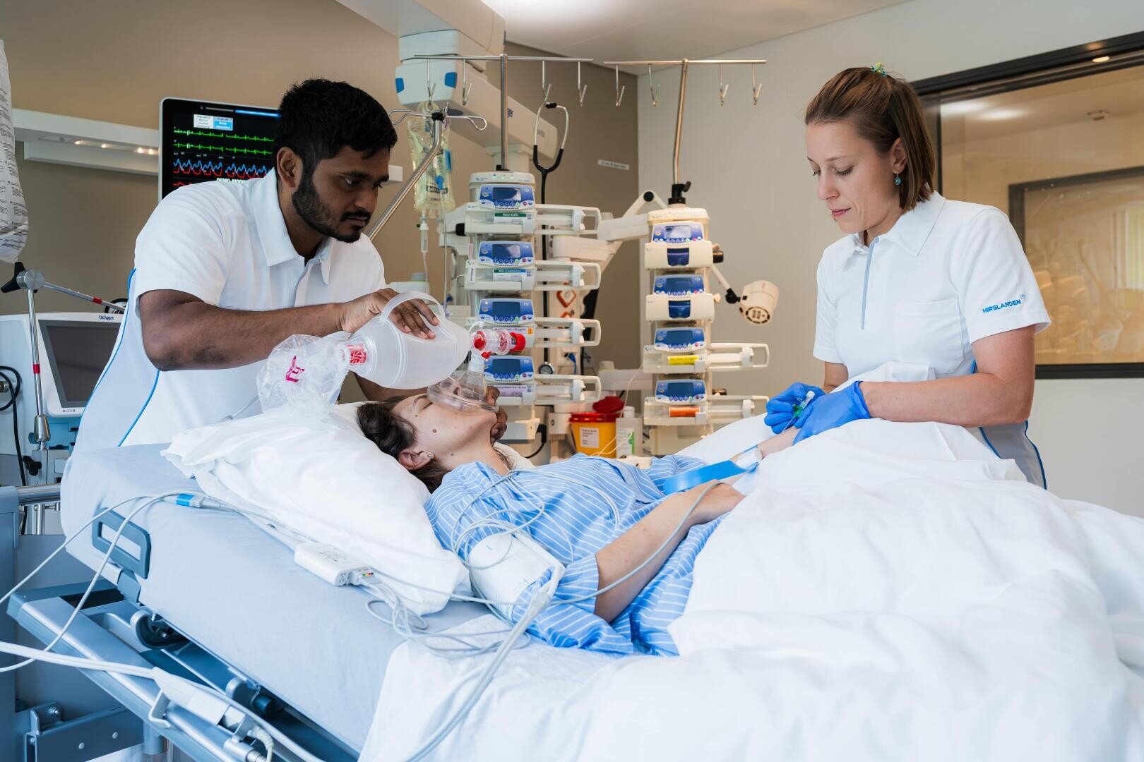 Ein Patient liegt in einem Krankenhausbett und wird versorgt, während eine männliche Pflegekraft Sauerstoff verabreicht und eine weibliche Pflegekraft die Geräte in einer Intensivstation überwacht. Medizinische Geräte und Monitore umgeben das Bett.