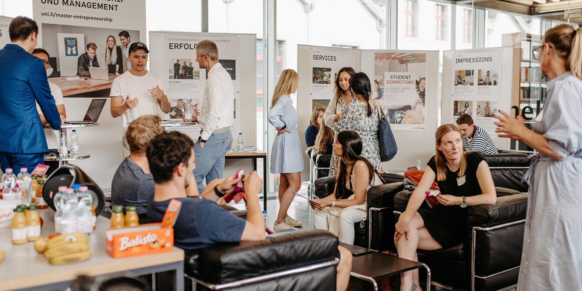Eine Gruppe von Menschen, die an einer Veranstaltung oder Ausstellung in einer Halle teilnehmen, unterhält sich, sitzt und läuft in einem modernen Raum mit Informationsplakaten an den Wänden und Erfrischungen auf einem Tisch herum.