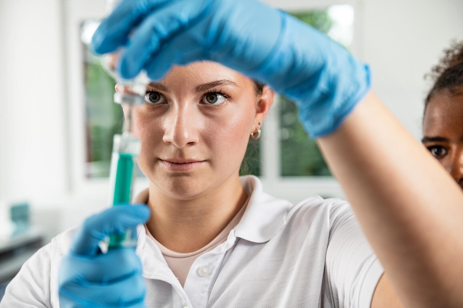 Eine Person, die blaue Handschuhe und ein weißes Hemd trägt, füllt in einer hellen Laborumgebung vorsichtig eine Spritze mit grüner Flüssigkeit aus einem Fläschchen.