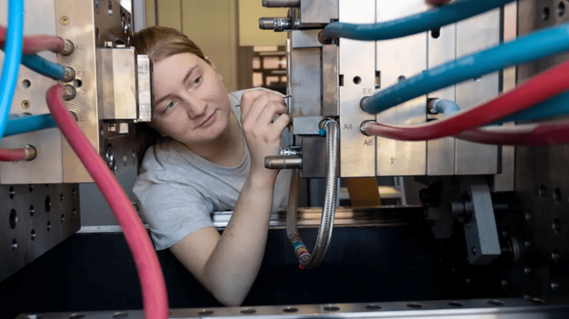 Eine Person in einem grauen Hemd arbeitet an Maschinen, justiert oder prüft Stecker und Kabel in einer industriellen Umgebung mit Metallteilen und bunten Röhren.