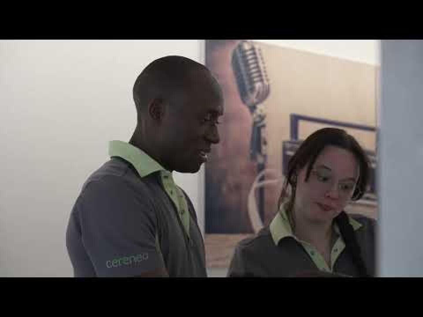 Zwei Personen in passenden grauen Hemden mit hellgrünen Kragen stehen in einem Haus, schauen etwas an, das nicht im Bild ist, und lächeln. Hinter ihnen hängt ein großes altes Mikrofonfoto an der Wand.