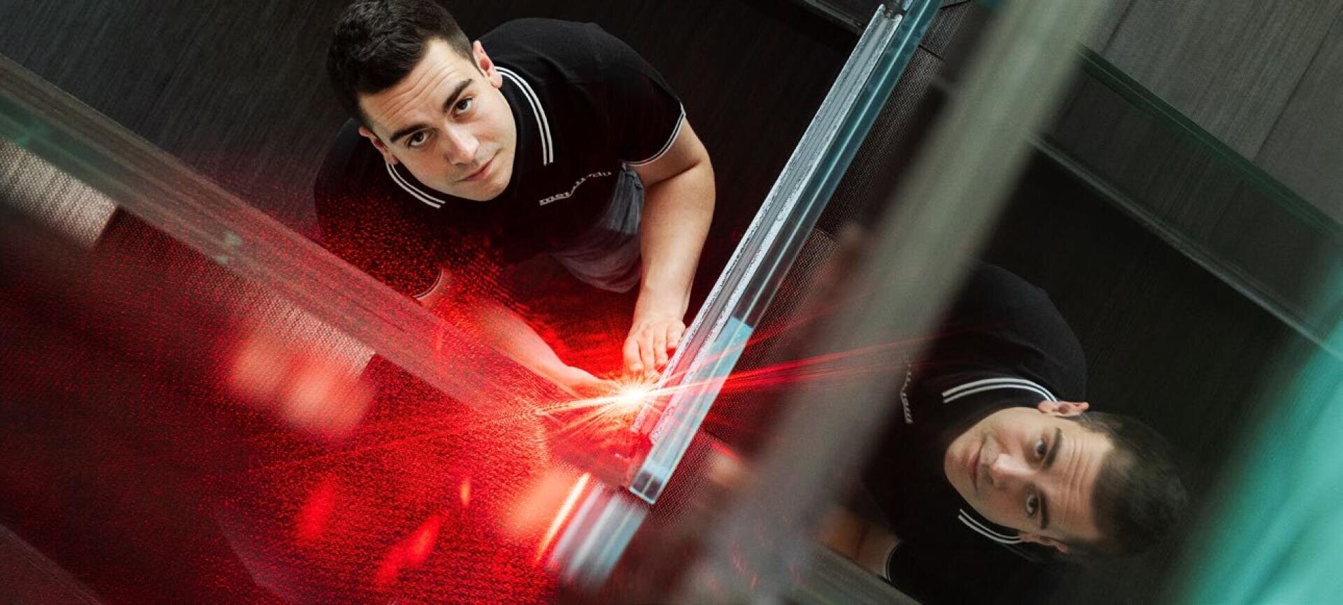 Eine Person in einem schwarzen Hemd hält ein Gerät, das einen roten Laserstrahl aussendet, und blickt in die Kamera, während sie in einem modernen, gläsernen Raum steht, in dem ihr Spiegelbild neben ihr zu sehen ist.