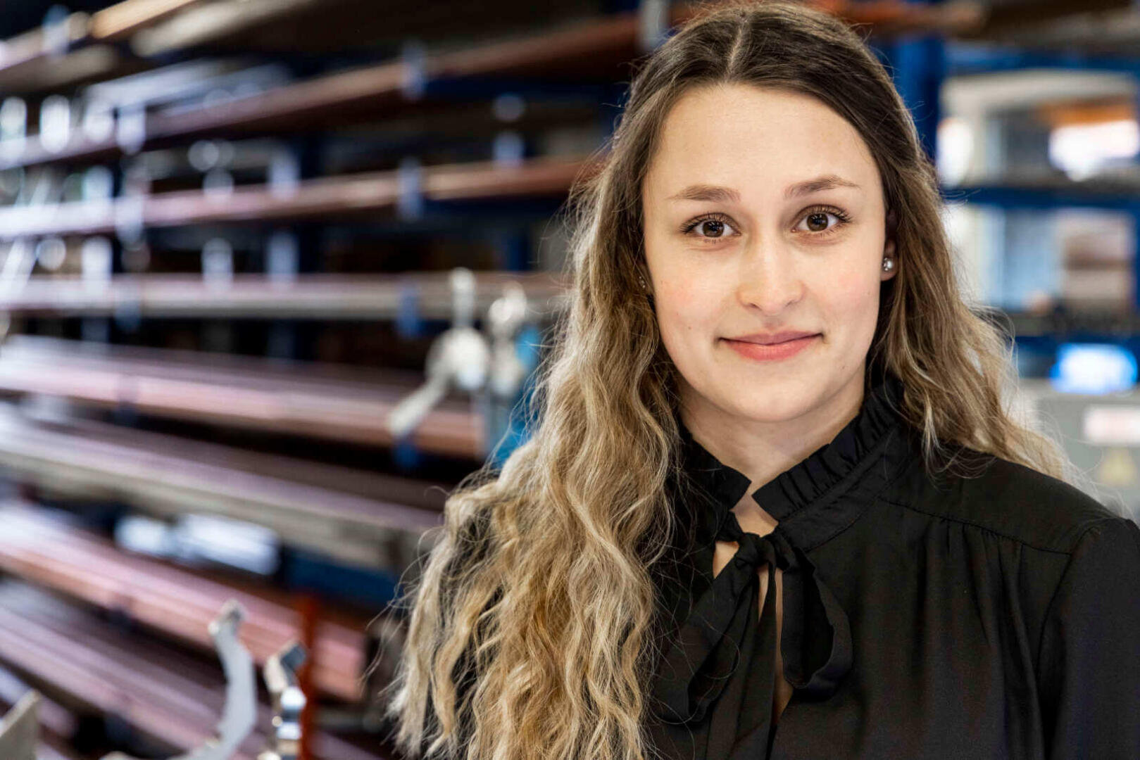 Eine Frau mit langen, gewellten Haaren, die eine schwarze Bluse trägt, steht in einem Innenraum und lächelt leicht. Im Hintergrund sind Regale mit aufgestapelten Metallstangen zu sehen, was auf eine Industrie- oder Werkstattaumgebung hindeutet.