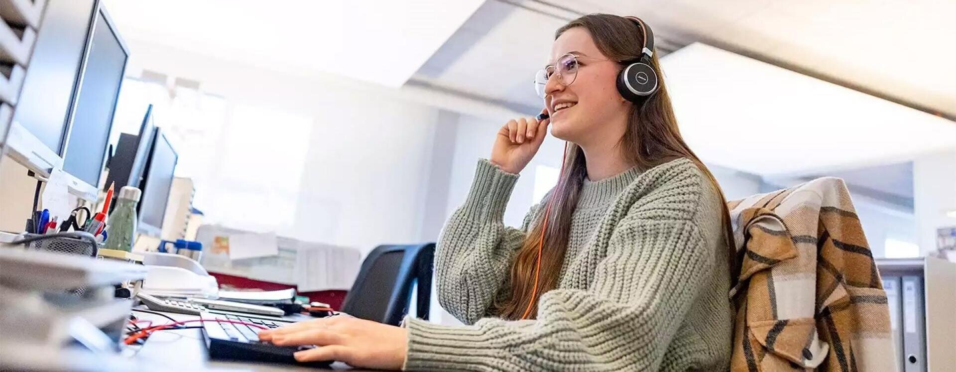 Eine junge Frau mit Brille und Kopfhörern sitzt an einem Schreibtisch in einem Büro und arbeitet lächelnd an einem Computer. Eine karierte Jacke ist über ihren Stuhl drapiert.
