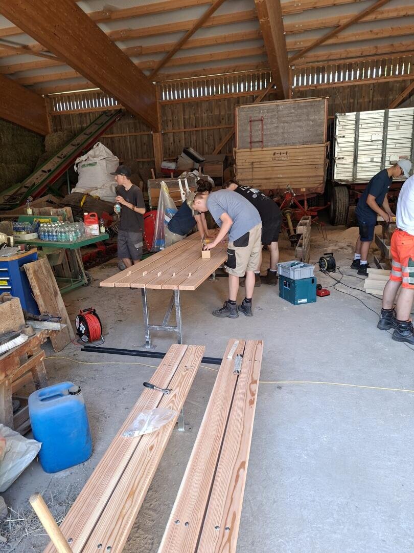 In einem großen Schuppen arbeiten mehrere Menschen zusammen und montieren Holzbretter auf Sägeböcken. Werkzeuge, Material und landwirtschaftliche Geräte liegen verstreut im Arbeitsbereich. Sonnenlicht strömt durch die offenen Wände und die Dachlatten herein.