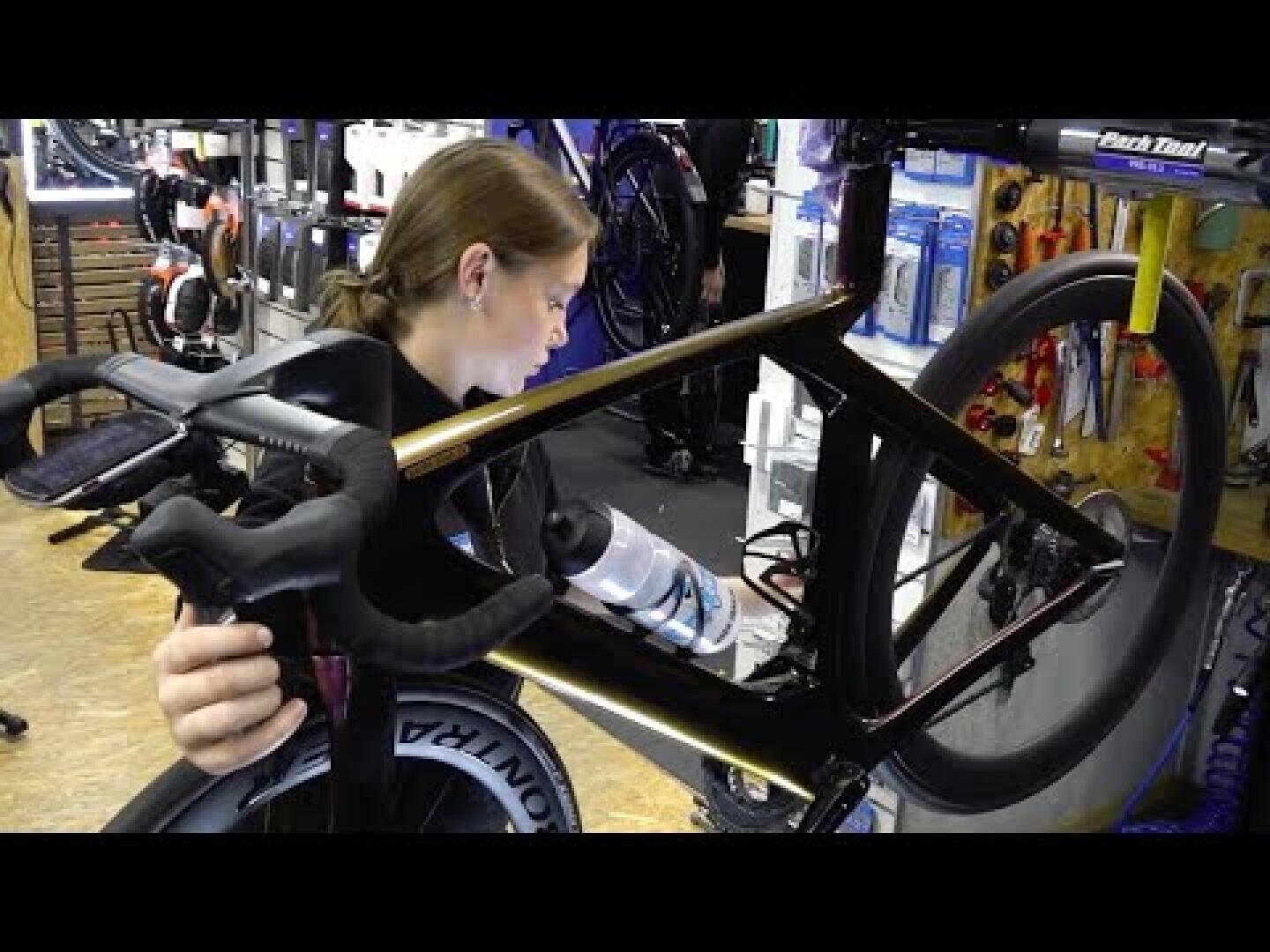 Eine Person arbeitet in einer Werkstatt an einem Rennrad und stellt die Komponenten in der Nähe des Rahmens ein. An der Wand im Hintergrund sind Werkzeuge und Fahrradzubehör zu sehen.