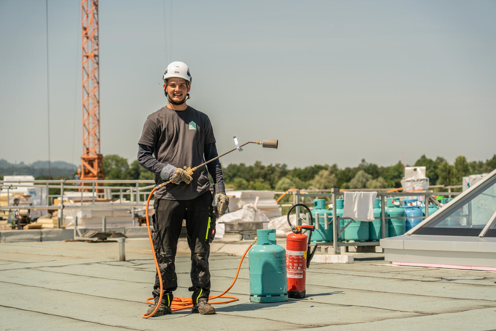 Ein Bauarbeiter mit Schutzhelm und Handschuhen steht auf einem Dach und hält eine Gasfackel, in der Nähe befinden sich Gasflaschen und ein Feuerlöscher. Im Hintergrund sind Baumaterialien und -geräte zu sehen.