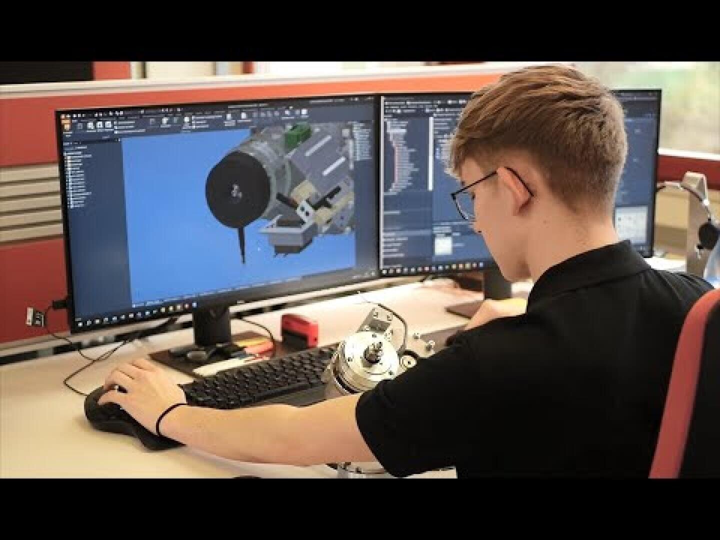 Eine Person sitzt an einem Schreibtisch mit einem Steuergerät vor zwei großen Monitoren, auf denen 3D-Modelle eines Raumfahrzeugs oder Satelliten in einem computergestützten Designprogramm angezeigt werden.