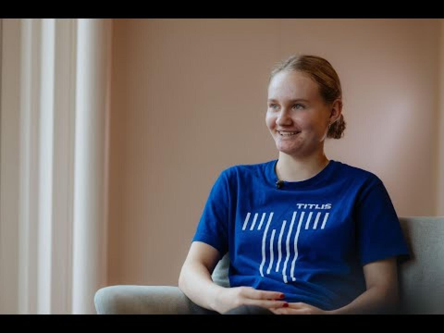 Eine Person mit hellen, nach hinten gebundenen Haaren und einem blauen TITLIS-T-Shirt sitzt auf einer hellen Couch und lächelt, wobei von links weiches, natürliches Licht einfällt.