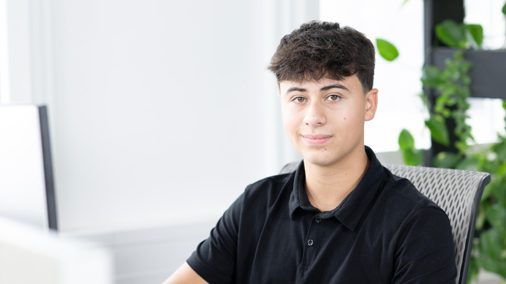 Ein junger Mann mit kurzen dunklen Haaren, der ein schwarzes Polohemd trägt, sitzt an einem Schreibtisch in einem hellen, modernen Büro mit Grünpflanzen im Hintergrund, schaut in die Kamera und lächelt leicht.