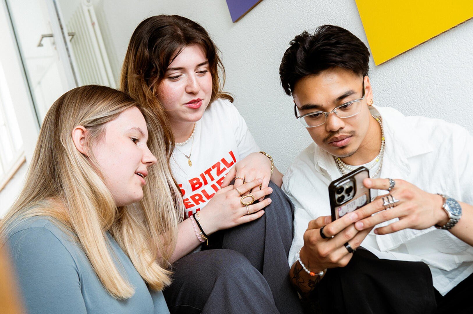 Drei junge Erwachsene sitzen eng beieinander und schauen aufmerksam auf ein Smartphone, das einer von ihnen in der Hand hält. Sie scheinen beschäftigt und konzentriert zu sein, möglicherweise diskutieren sie oder schauen sich etwas auf dem Telefon an.