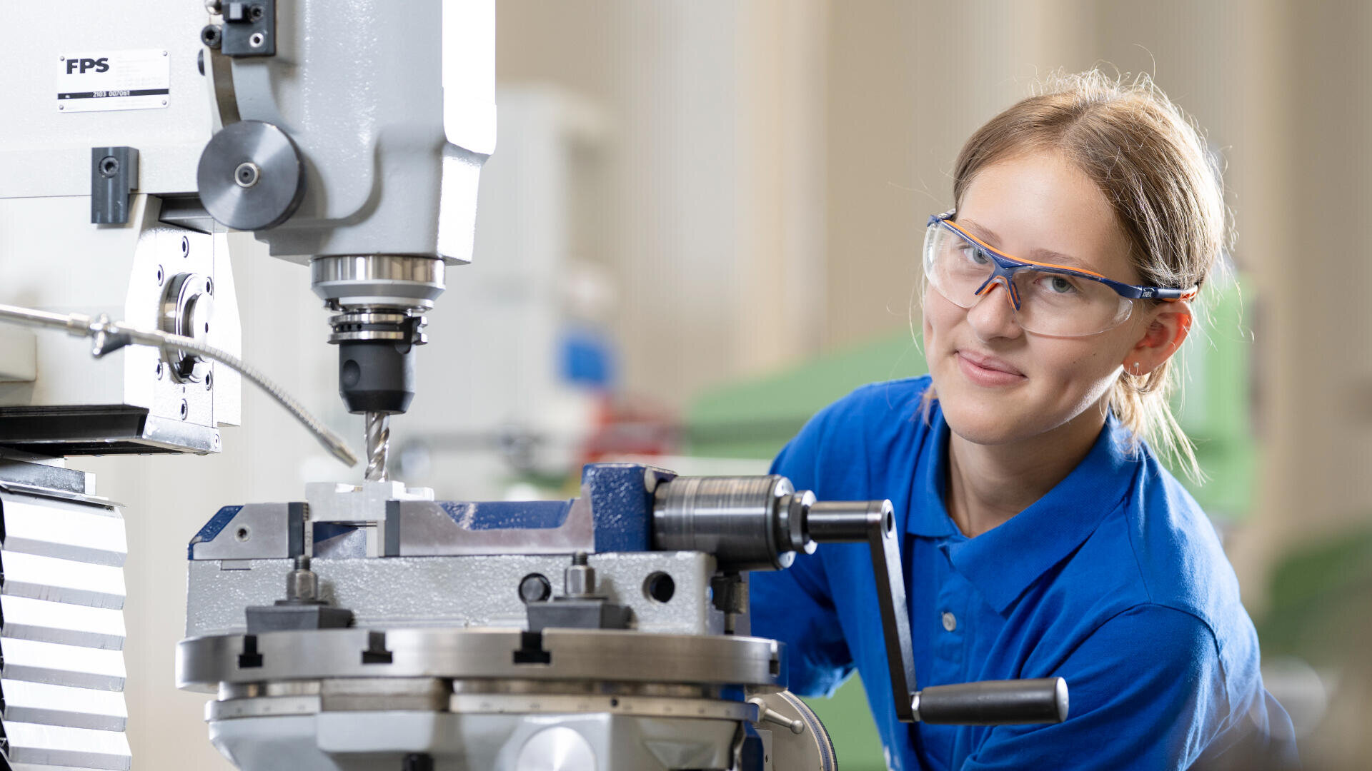 Eine Person mit Schutzbrille und blauem Hemd bedient eine Fräsmaschine in einer Werkstatt und konzentriert sich auf die Metallbearbeitung.