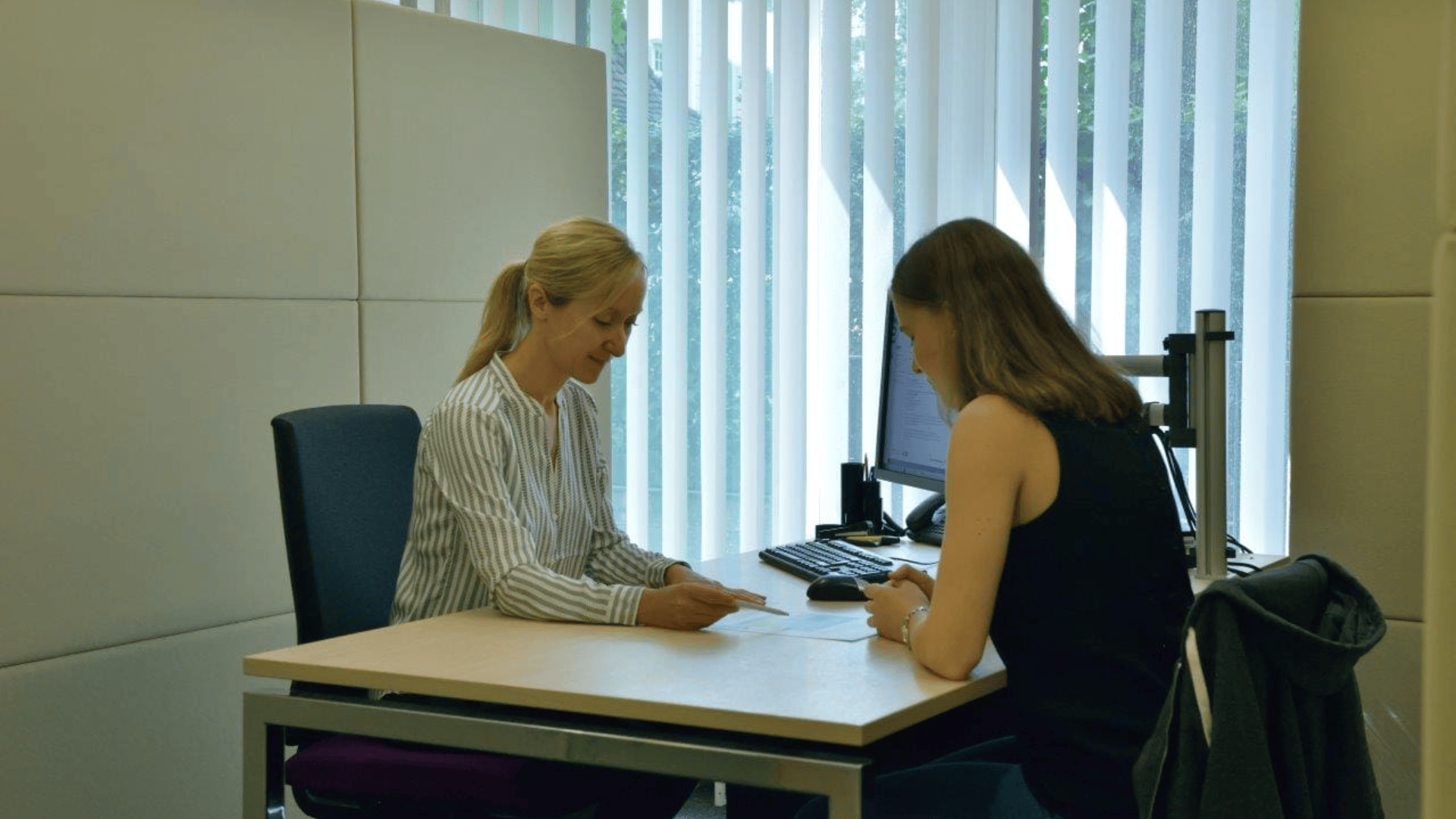 Zwei Frauen sitzen sich an einem Bürotisch gegenüber. Die eine reicht der anderen ein Papier. Ein Computer und Büromaterialien sind zu sehen, vertikale Jalousien lassen natürliches Licht durch ein Fenster hinter ihnen herein.