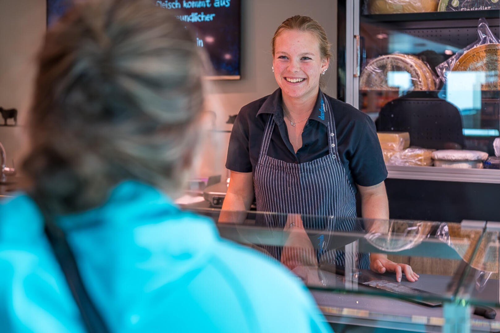Ein lächelnder Mitarbeiter eines Feinkostladens, der eine Schürze trägt und hinter einer Glastheke steht, bedient einen Kunden.