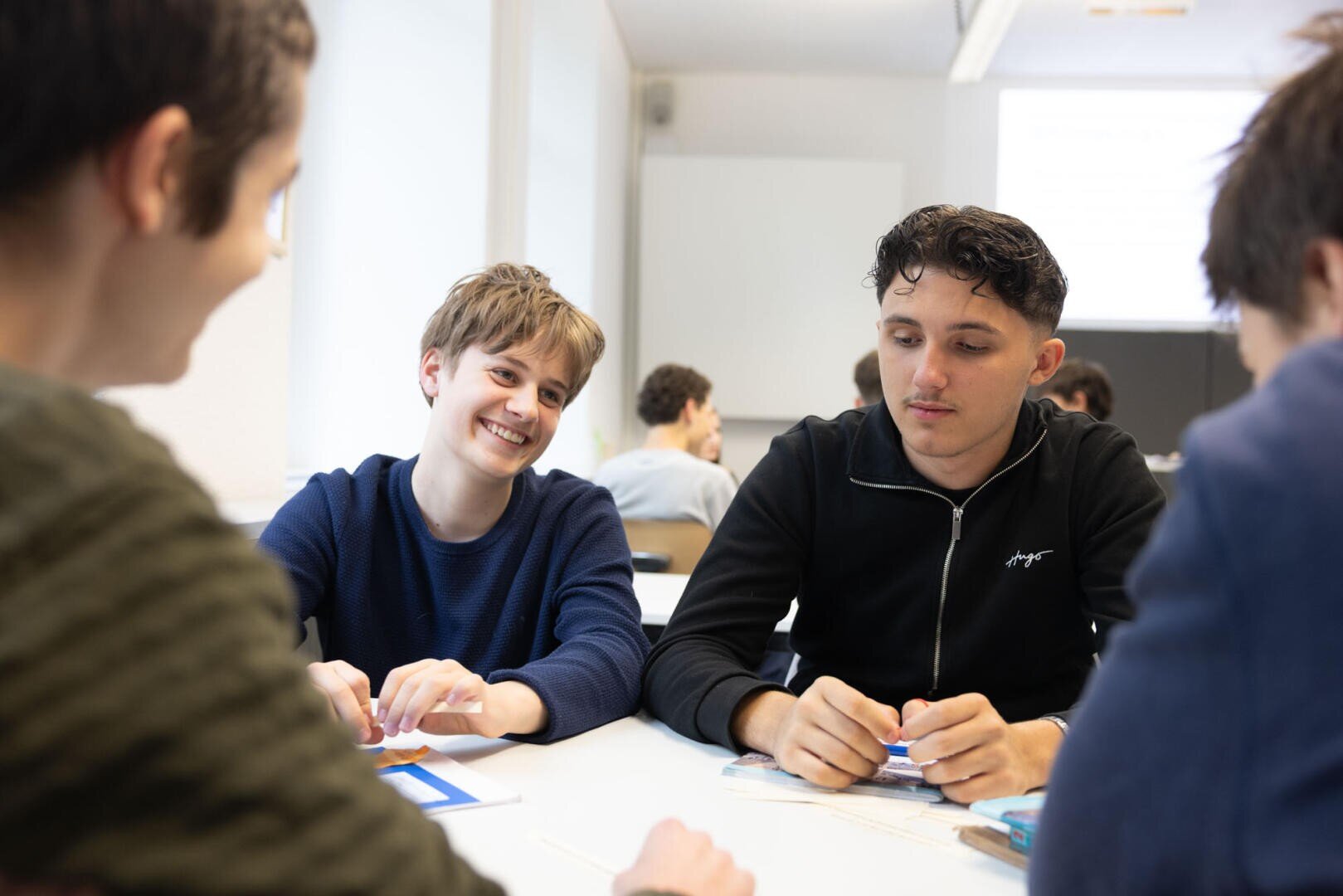 Vier junge Erwachsene sitzen um einen Tisch in einem hellen Klassenzimmer und unterhalten sich. Zwei von ihnen lächeln, und auf dem Tisch liegen Papiere und Stifte, was darauf hindeutet, dass sie an einer Gruppenaktivität oder Diskussion arbeiten.