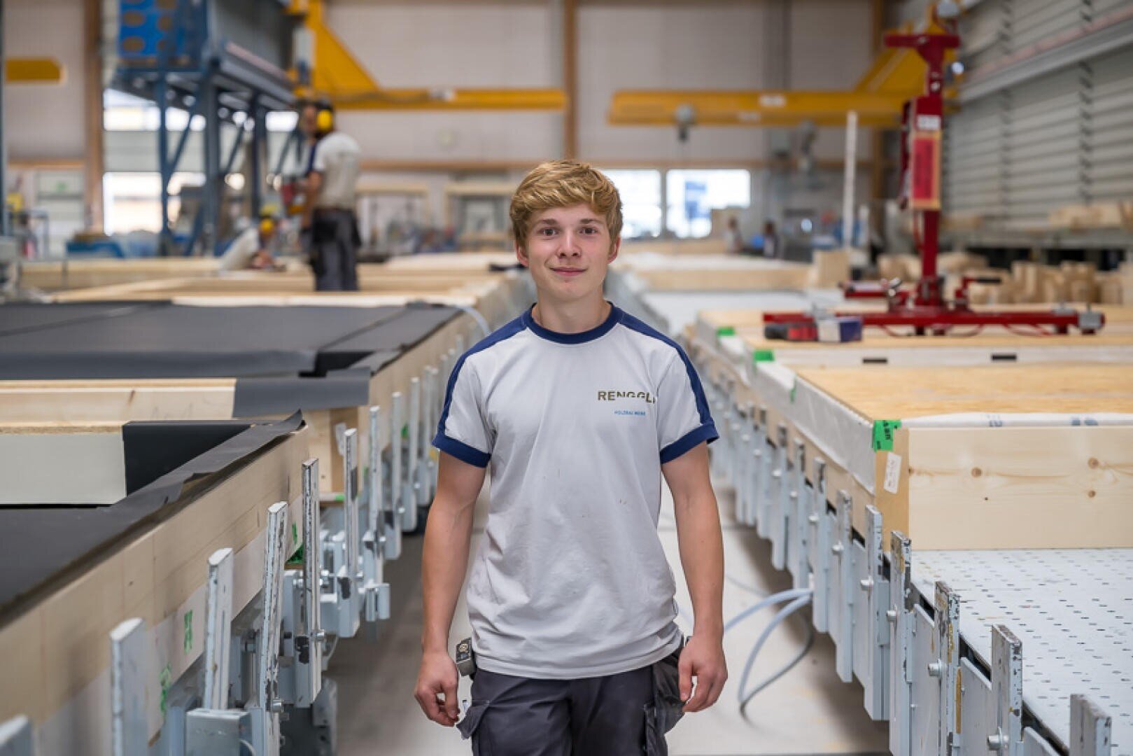 Ein junger Mann in einem weiß-blauen T-Shirt steht lächelnd in einer modernen Fabrik oder Werkstatt, umgeben von Industrieanlagen und Holzmaterialien, mit einem anderen Arbeiter im Hintergrund.