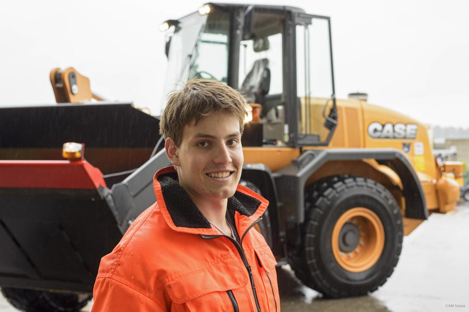 Ein junger Mensch in einer orangefarbenen Warnweste lächelt an einem bewölkten Tag vor einem großen gelben CASE-Baufahrzeug.