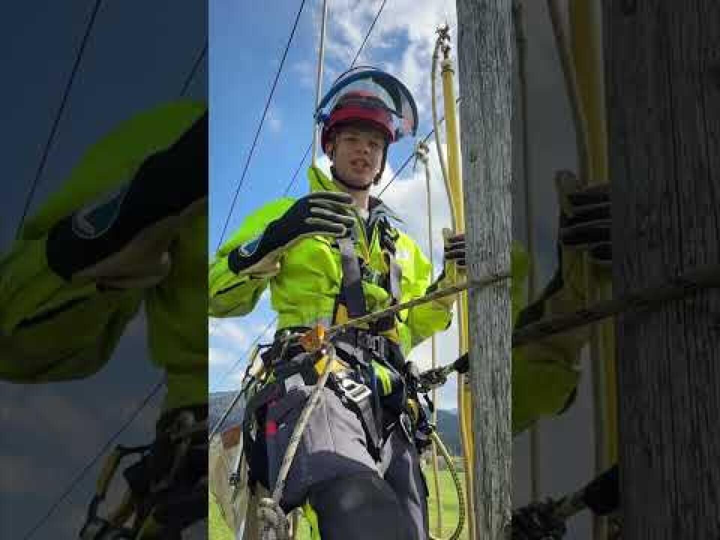 Ein Versorgungsarbeiter in Schutzkleidung und Helm steht auf einer Leiter neben einem hölzernen Versorgungsmast und hält eine Ausrüstung. Im Hintergrund sind Stromleitungen und blauer Himmel zu sehen.