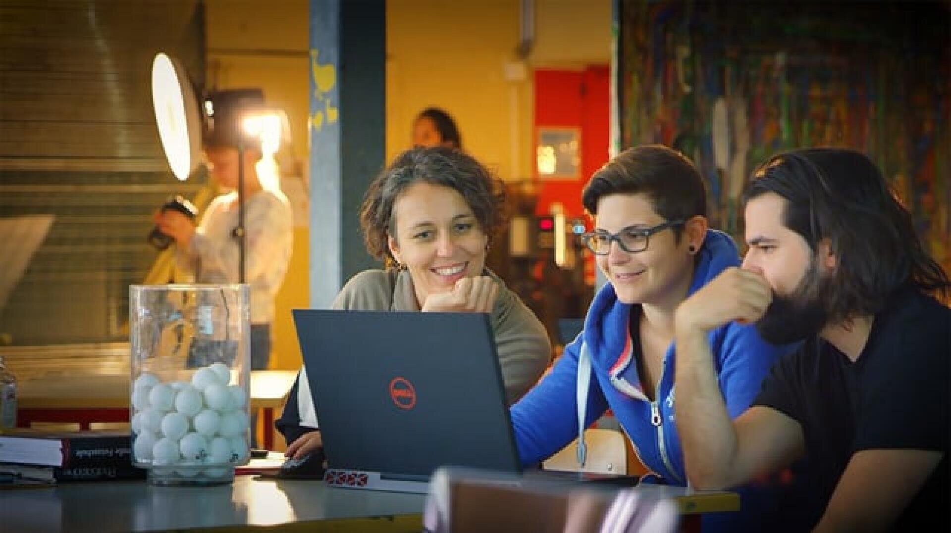 Drei Personen sitzen an einem Tisch, lächeln und schauen auf einen Laptop-Bildschirm. Auf dem Tisch steht ein großes Glas mit Tischtennisbällen. Der Hintergrund ist hell beleuchtet, mit bunten Dekorationen geschmückt, und eine weitere Person steht dort.