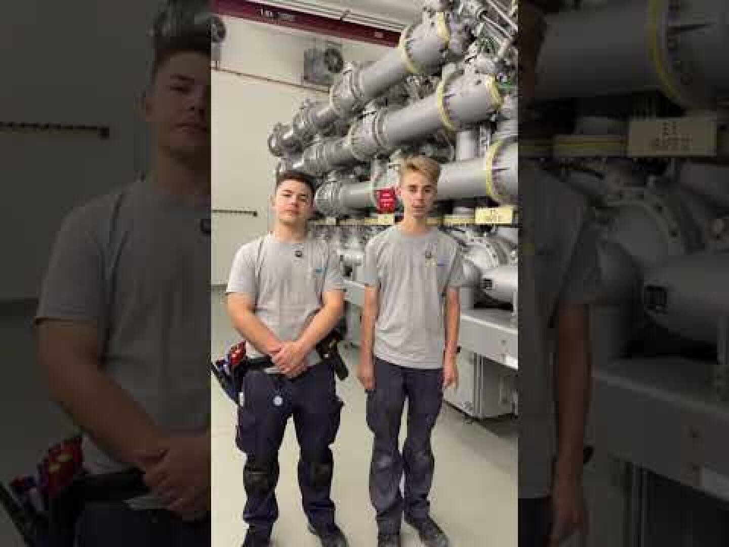 Zwei junge Männer in grauen Hemden und dunklen Hosen stehen nebeneinander vor einer großen Industrieanlage mit Rohren und Ventilen, bei der es sich anscheinend um eine Industrie- oder Versorgungseinrichtung handelt.