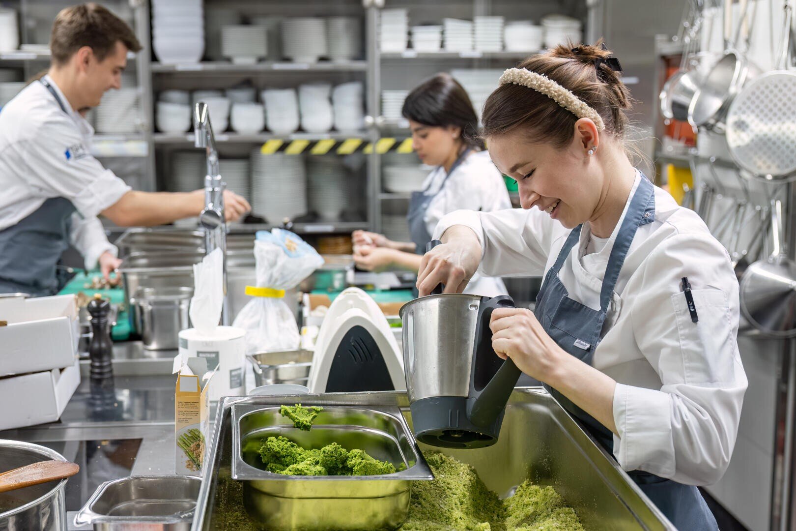 Drei Köche arbeiten in einer geschäftigen Küche. Im Vordergrund schüttet ein lächelnder Koch im weißen Kittel grüne Soße aus einem Mixer in ein Metalltablett. Im Hintergrund hängen Regale mit Tellern und Utensilien.
