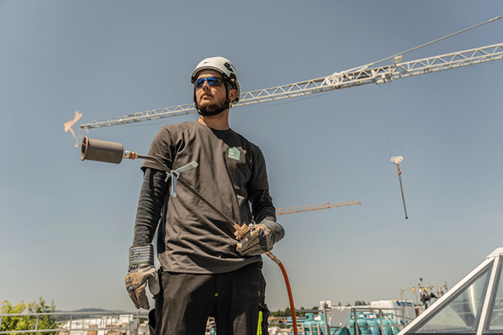 Ein Bauarbeiter in Schutzkleidung hält auf einer Baustelle einen brennenden Schweißbrenner in der Hand; im Hintergrund sind unter strahlend blauem Himmel Kräne und Baumaschinen zu sehen.