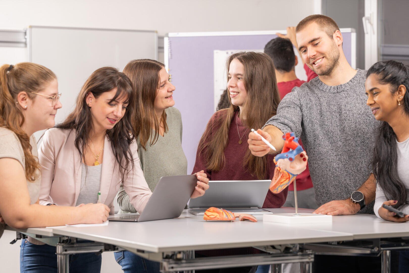 Eine Gruppe von fünf jungen Erwachsenen steht um einen Tisch mit Laptops, Papieren und anatomischen Modellen, darunter ein Herz. Sie lächeln und sind in eine Diskussion vertieft, was auf eine gemeinschaftliche Lernumgebung hindeutet.
