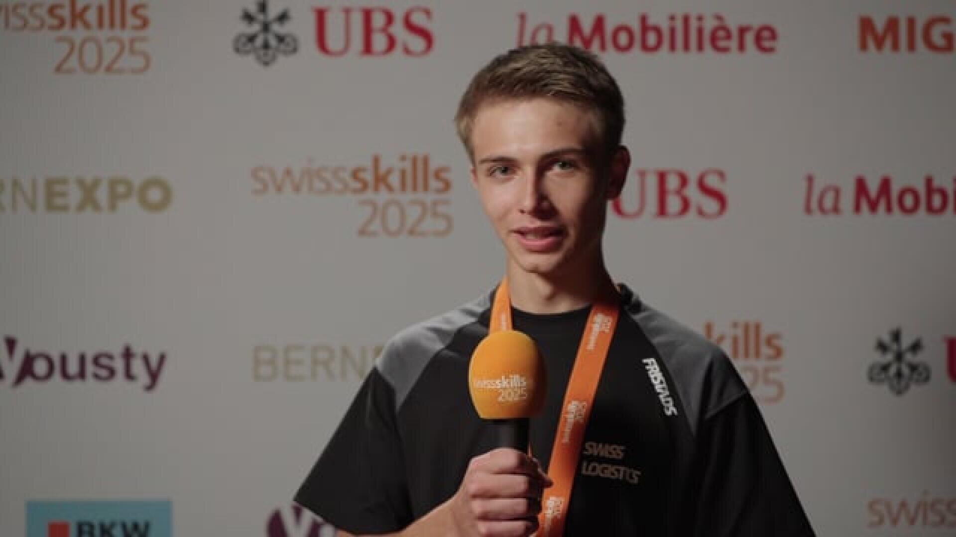 Ein junger Mann mit Swiss Logistics-Shirt und orangefarbenem Medaillenband spricht in ein orangefarbenes Mikrofon mit der Aufschrift SwissSkills 2025 und steht vor einer Kulisse mit verschiedenen Sponsorenlogos.