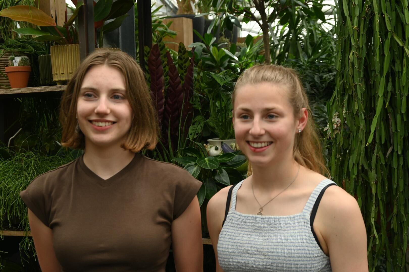 Zwei junge Frauen, die lächelnd nebeneinander in einem üppigen Innengarten stehen, im Hintergrund sind verschiedene Grünpflanzen zu sehen.