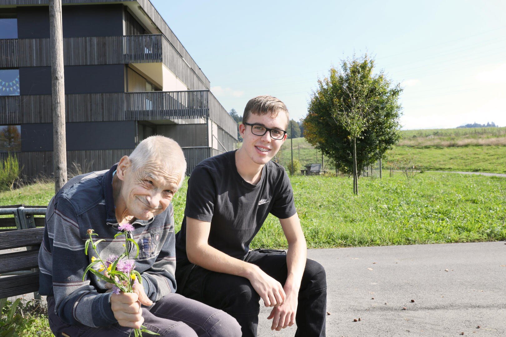 Ein Mitarbeitender (ehemals Lernender FaBe) sitzt zusammen mit einem Klienten auf einer Bank bei Sonnenschein.