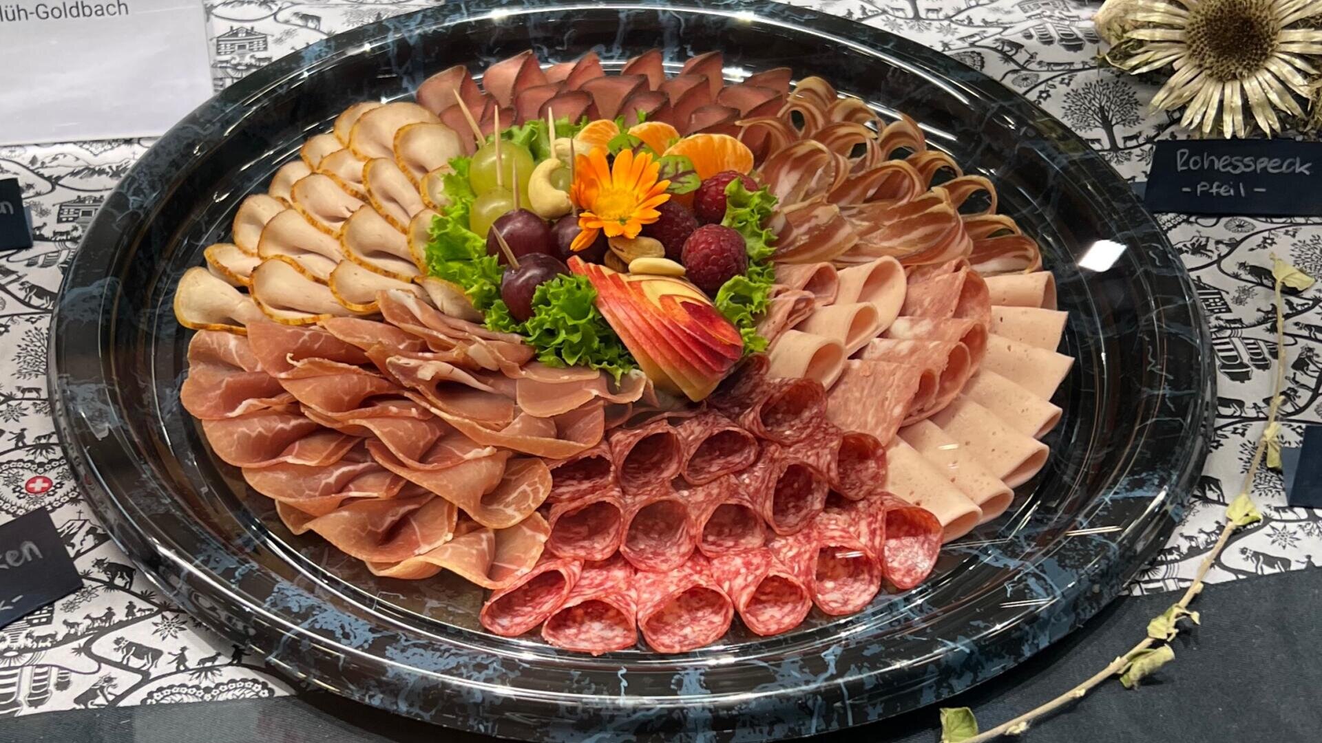 Eine Platte mit ordentlich angeordneten Aufschnittscheiben, darunter Salami, Schinken und Putenbrust, garniert mit Salat, Weintrauben, einem in Scheiben geschnittenen Apfel und einer essbaren Blüte, serviert auf einem Marmortablett.