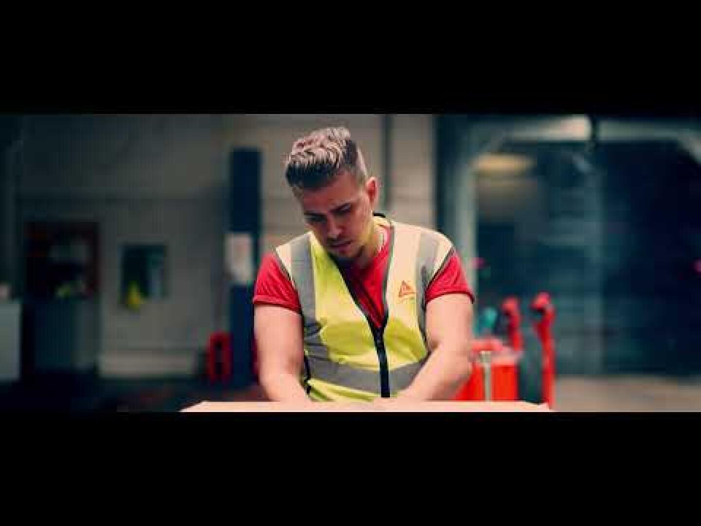 Ein Mann, der eine gelbe Sicherheitsweste und ein rotes Hemd trägt, steht in einem Gebäude und arbeitet konzentriert an einem Tisch oder verpackt eine Kiste in einer Industrie- oder Lagerhalle.