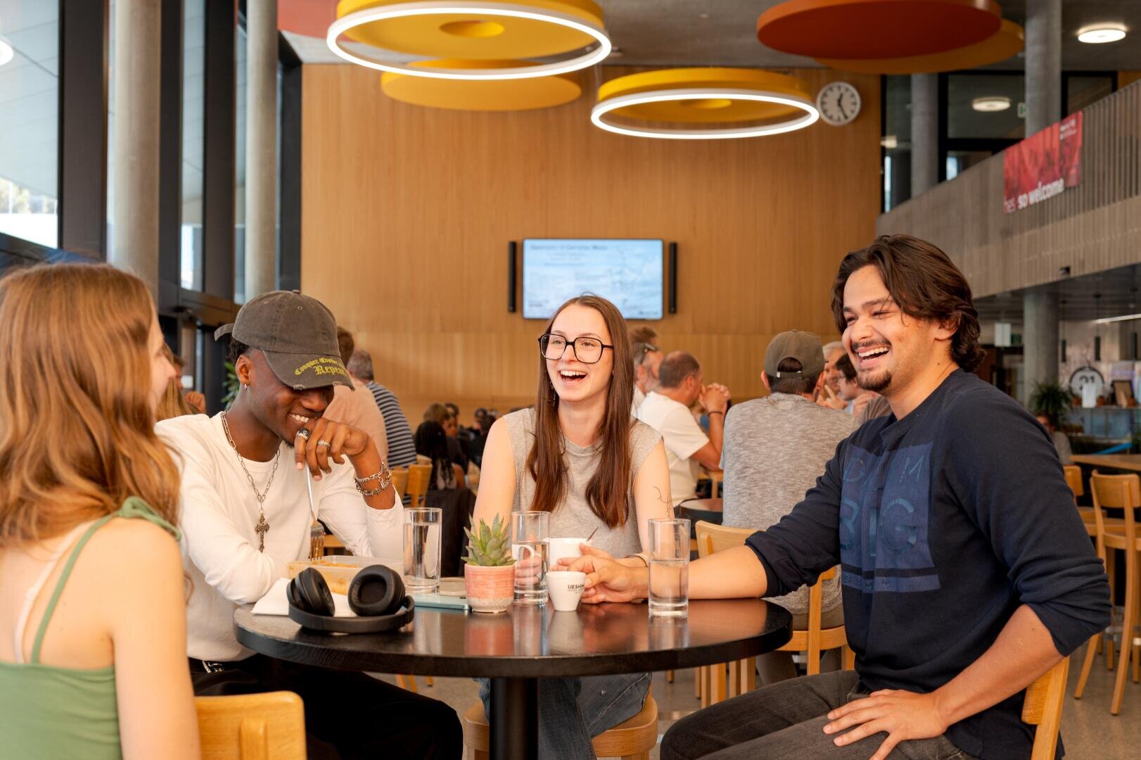 Vier junge Erwachsene sitzen in einem modernen Café um einen Tisch herum, lachen und unterhalten sich. Vor sich haben sie Getränke stehen und scheinen die gemeinsame Zeit zu genießen. Im Hintergrund sitzen weitere Gäste an Tischen.