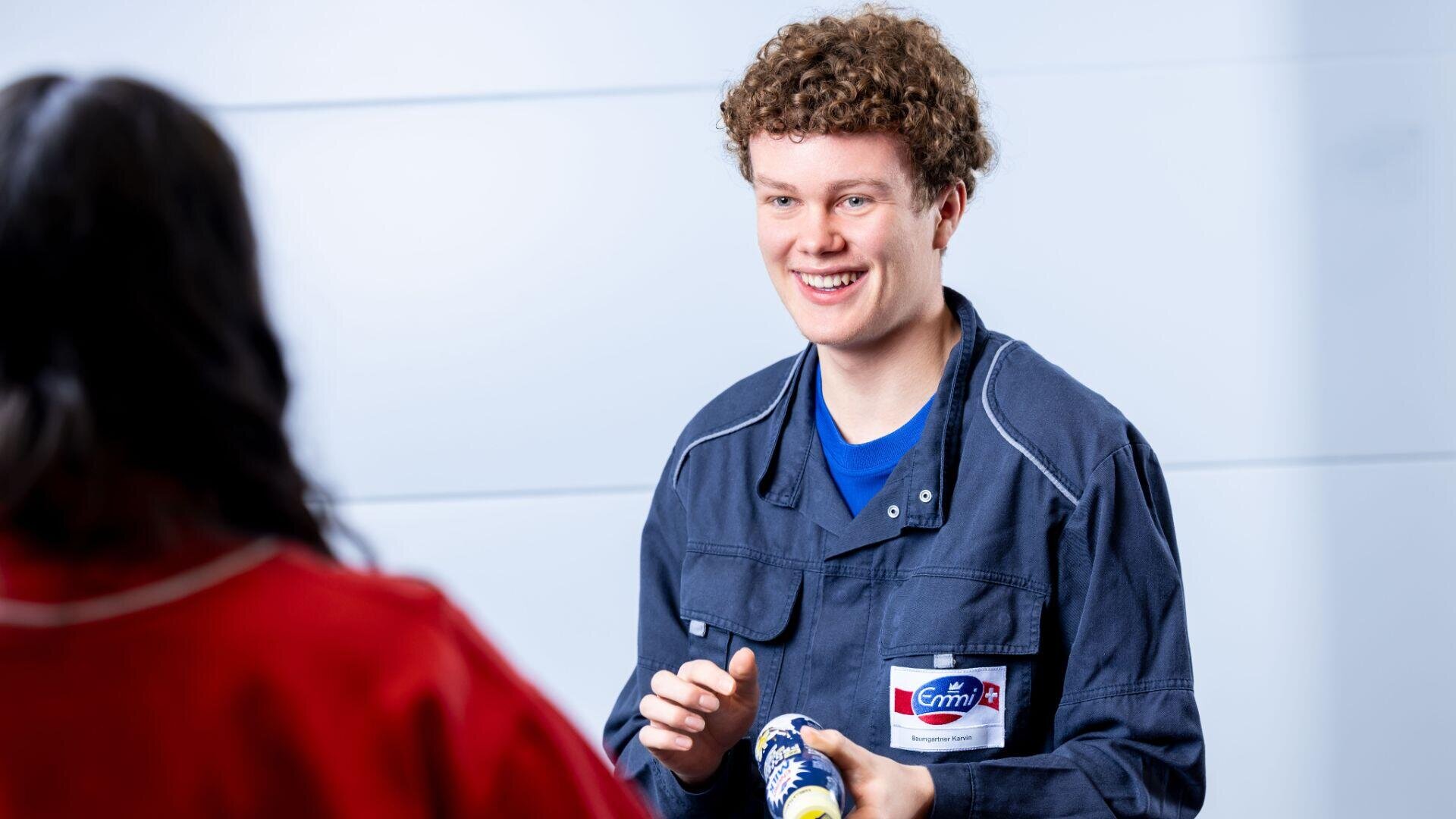 Eine Person in einer blauen Uniform mit Namensschild lächelt, während sie einen verpackten Gegenstand in der Hand hält, und blickt dabei einer anderen Person in roter Kleidung entgegen. Der Hintergrund ist unscharf und hell.