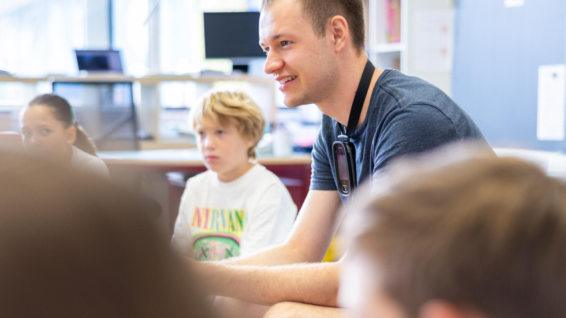 Ein junger Erwachsener, der ein Mikrofon-Headset trägt, sitzt in einem hell erleuchteten Klassenzimmer, lächelt und unterhält sich mit Kindern, die in der Nähe sitzen. Im Hintergrund sind Computermonitore und Regale zu sehen.