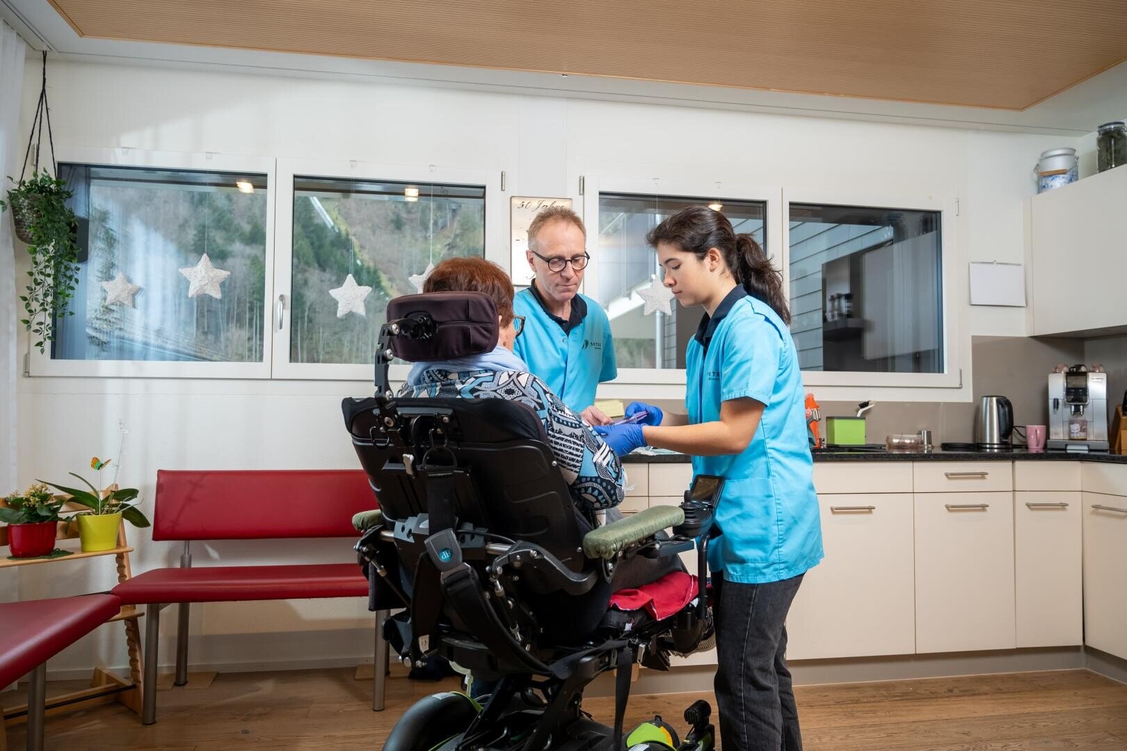 Zwei Pflegekräfte in blauen Uniformen helfen einer Person im Rollstuhl in einem hellen, modernen Raum mit großen Fenstern, Topfpflanzen und Küchengeräten im Hintergrund.