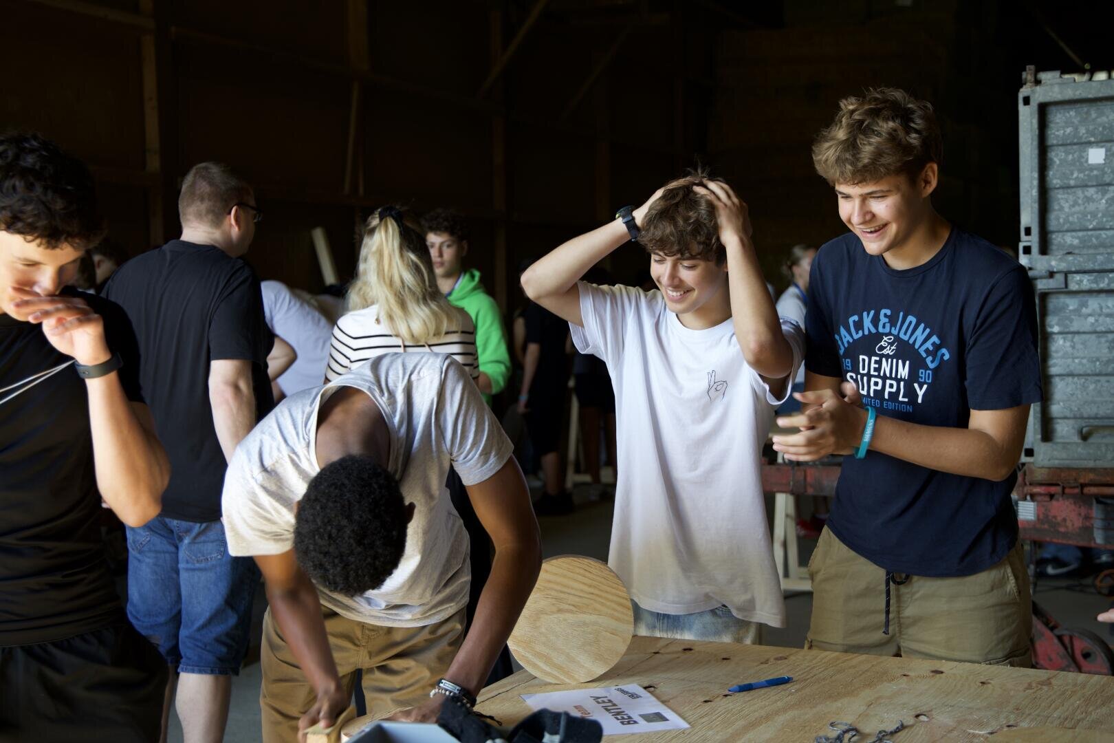 Eine Gruppe Jugendlicher versammelt sich um einen Holztisch, einige lächeln und lachen, während andere konzentriert an einem Projekt zu arbeiten scheinen. Sie befinden sich in einer schwach beleuchteten Werkstatt oder Garage, in der Werkzeuge und Materialien auf dem Tisch liegen.