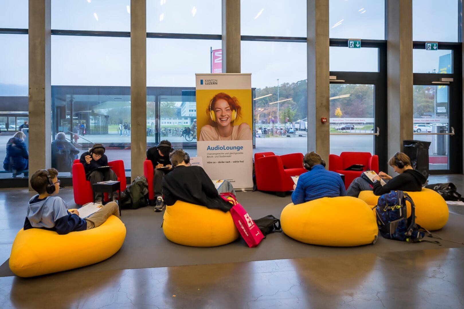Mehrere Personen sitzen auf leuchtend gelben Sitzsäcken und roten Stühlen und hören mit Kopfhörern in einem modernen Lounge-Bereich im Innenbereich Audio. Im Hintergrund wirbt ein großes Banner mit einer lächelnden Person für AudioLounge.
