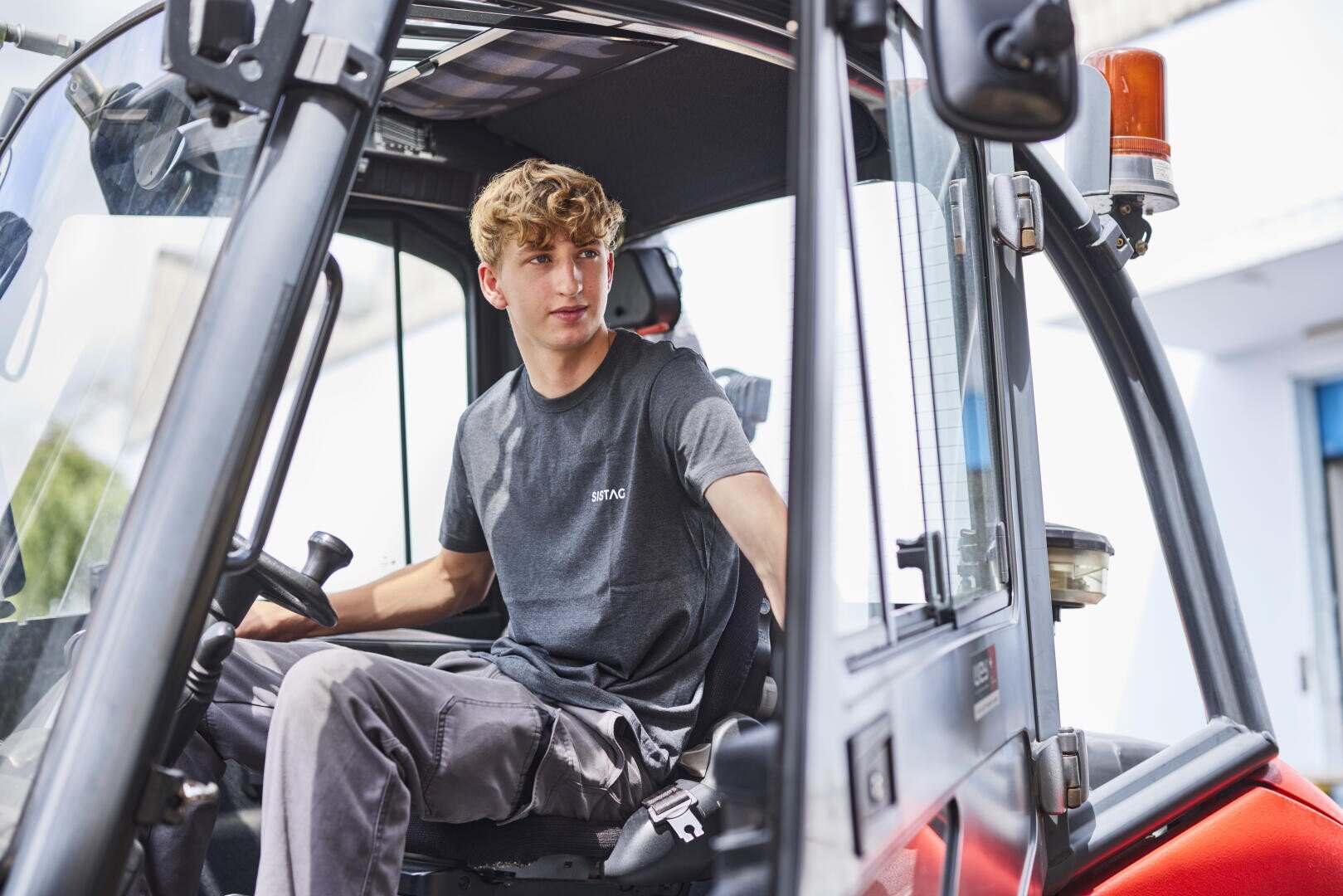 Ein junger Mann mit hellbraunem Haar sitzt auf dem Fahrersitz eines Gabelstaplers und schaut nach hinten, während er die Bedienelemente bedient. Er trägt ein graues T-Shirt und eine graue Hose in einer hellen, industriellen Umgebung.