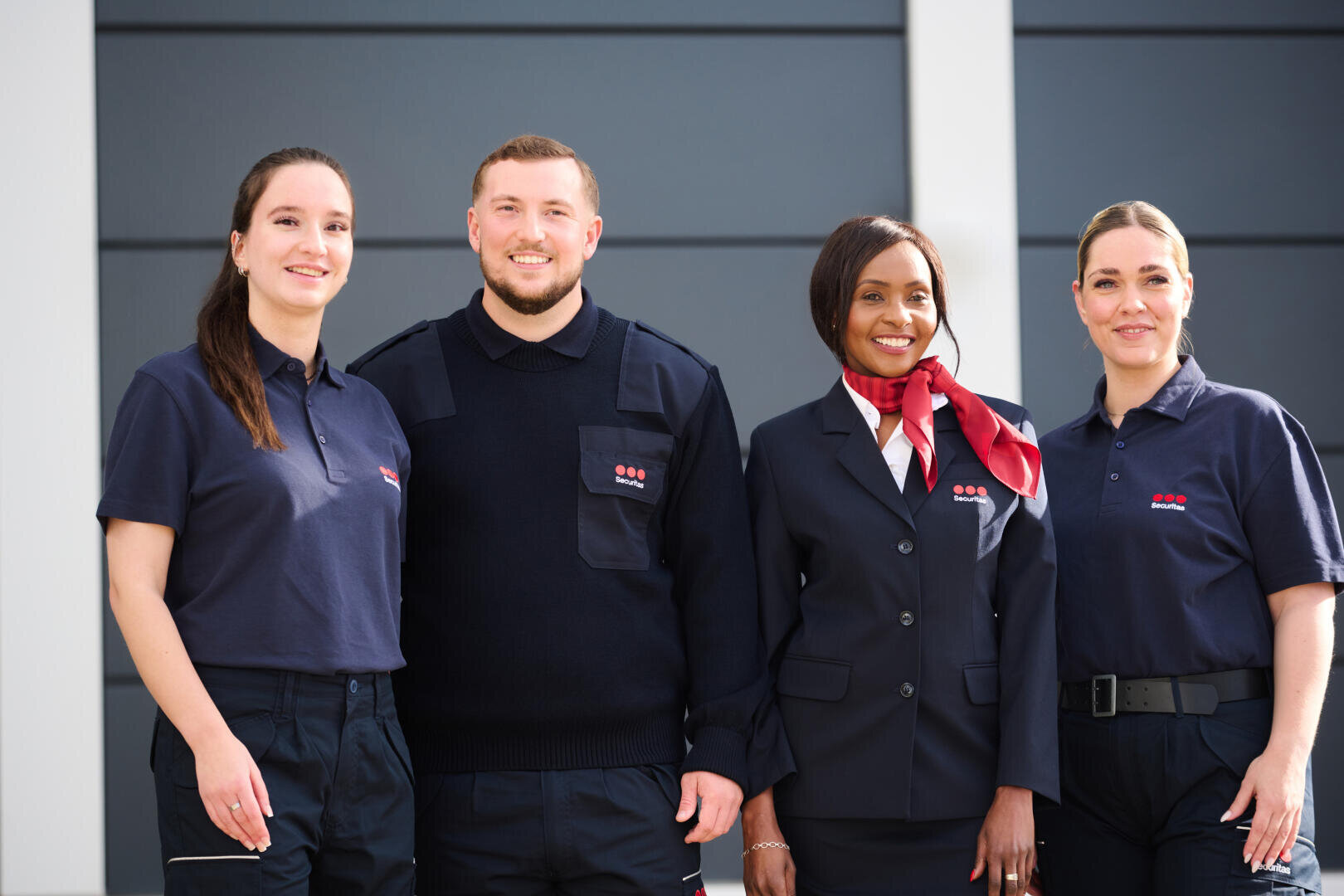 Vier Sicherheitsfachkräfte – drei Frauen und ein Mann – stehen lächelnd in Uniform vor einem modernen Gebäude mit grauen Fassadenplatten.